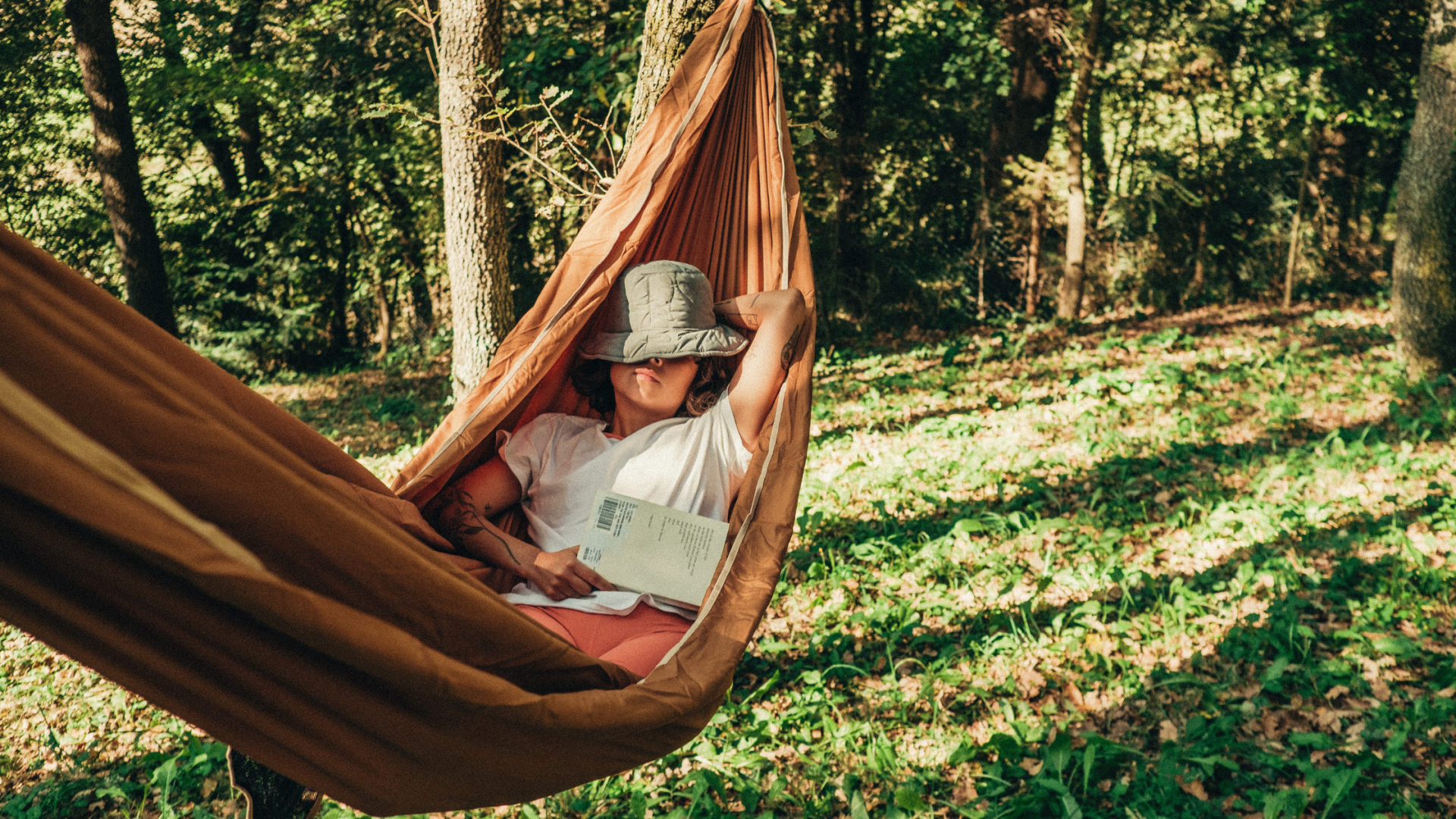 a person relaxing on a hammock outdoor with a hat covering their face while hokding a book