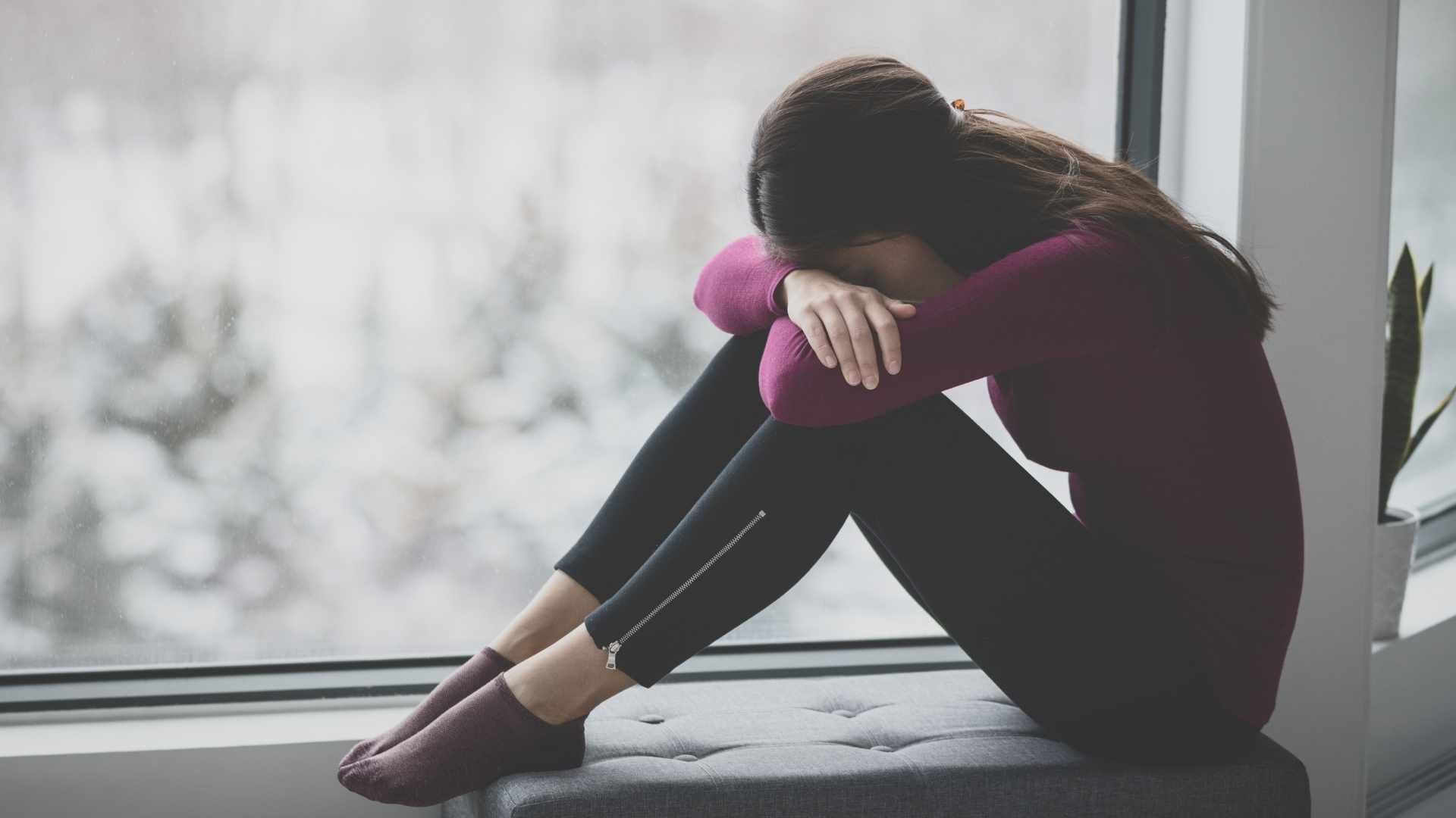 woman with her head down on her arms and knees beside the window feeling sad