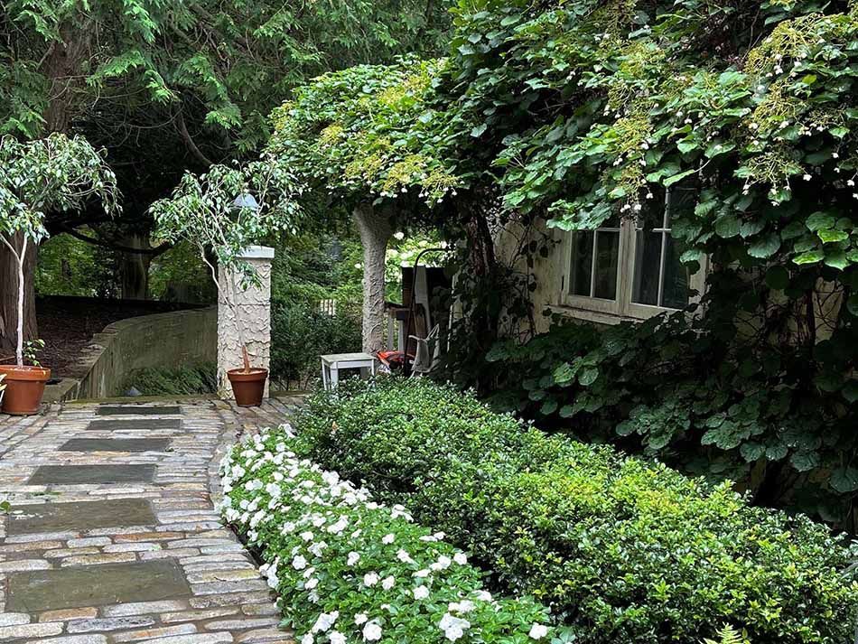 Stone path through a lush garden with bushes, flowers, and a covered porch.