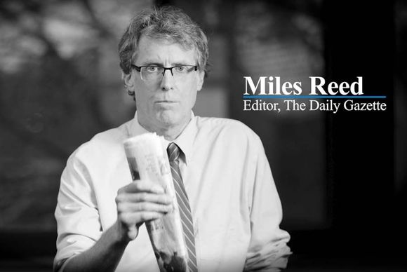 Miles Reed, editor of The Daily Gazette, holding a newspaper. Black and white photo with serious expression.