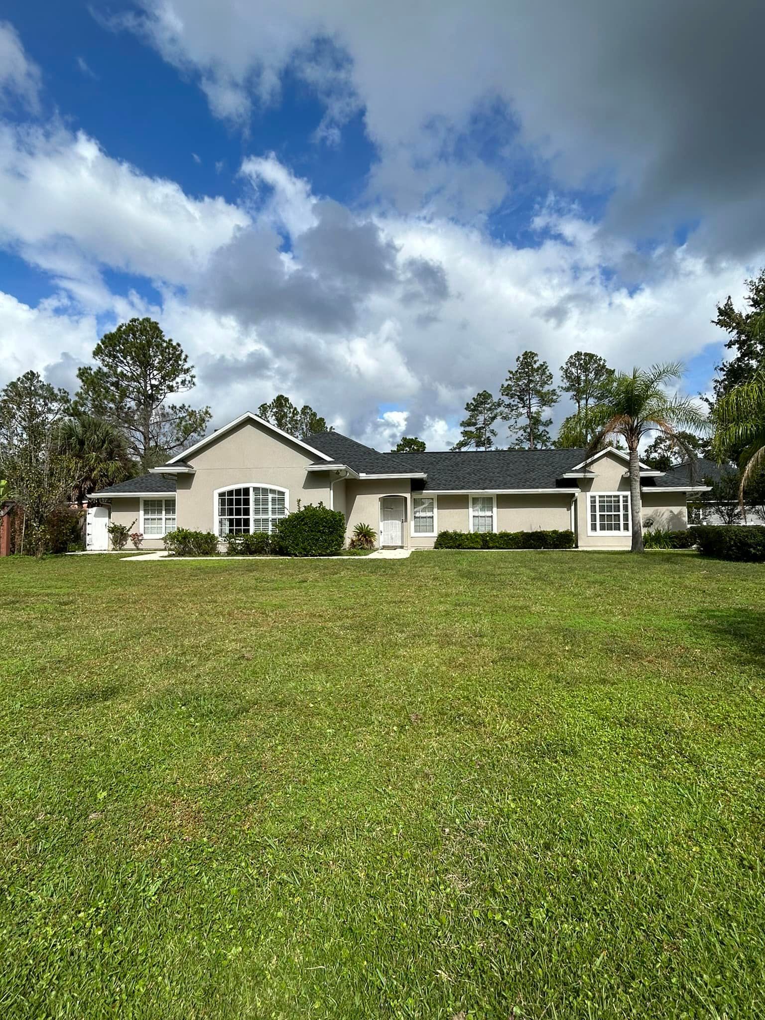 Beige ranch-style house with dark roof and green lawn on a partly cloudy day.