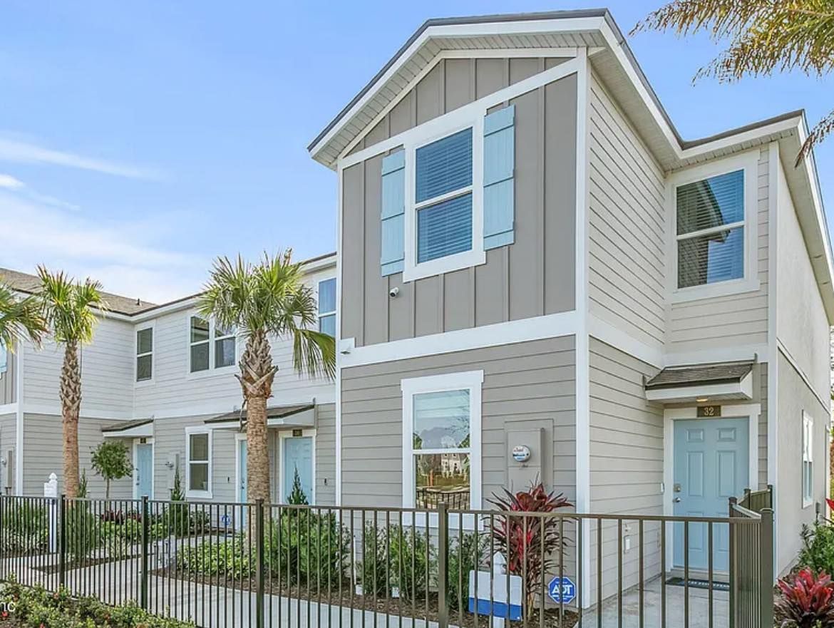 Two-story beige townhouses with blue doors and shutters, behind a black fence, with palm trees.