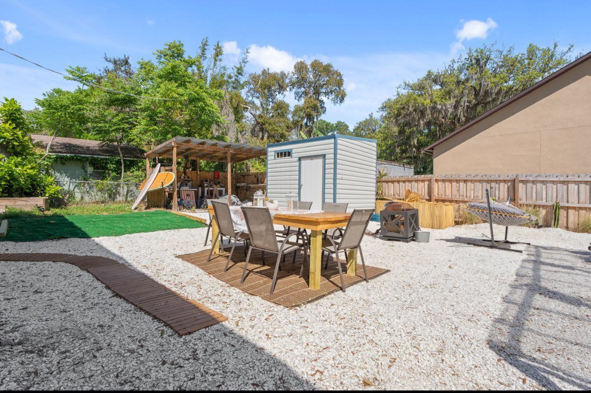 Backyard with a dining table on gravel, shed, covered storage area, and trees.