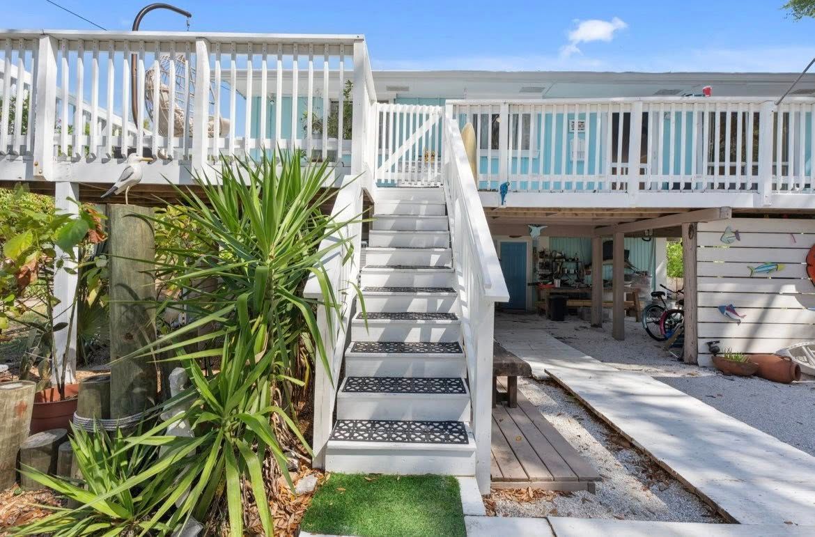White beach house with elevated deck and stairs, blue accents, and lush tropical plants.