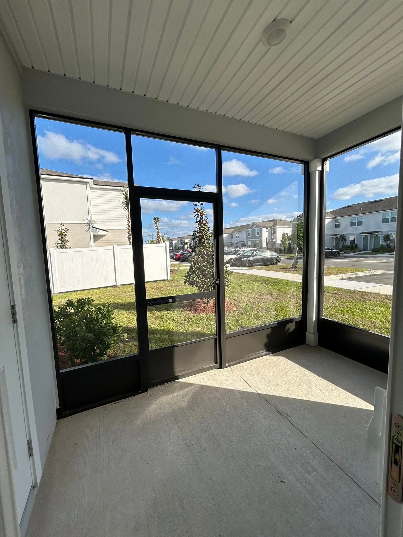 Screened porch with dark frames and concrete floor, view of a yard and houses on a sunny day.