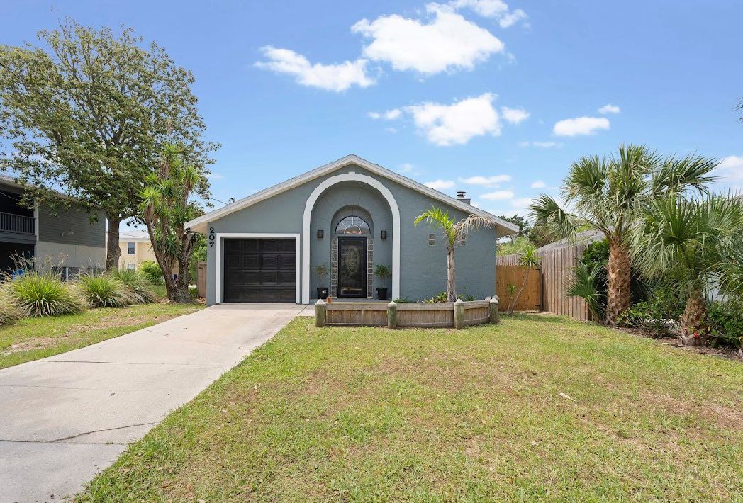 Blue house with arched front entry, driveway, and patchy grass under a blue sky.