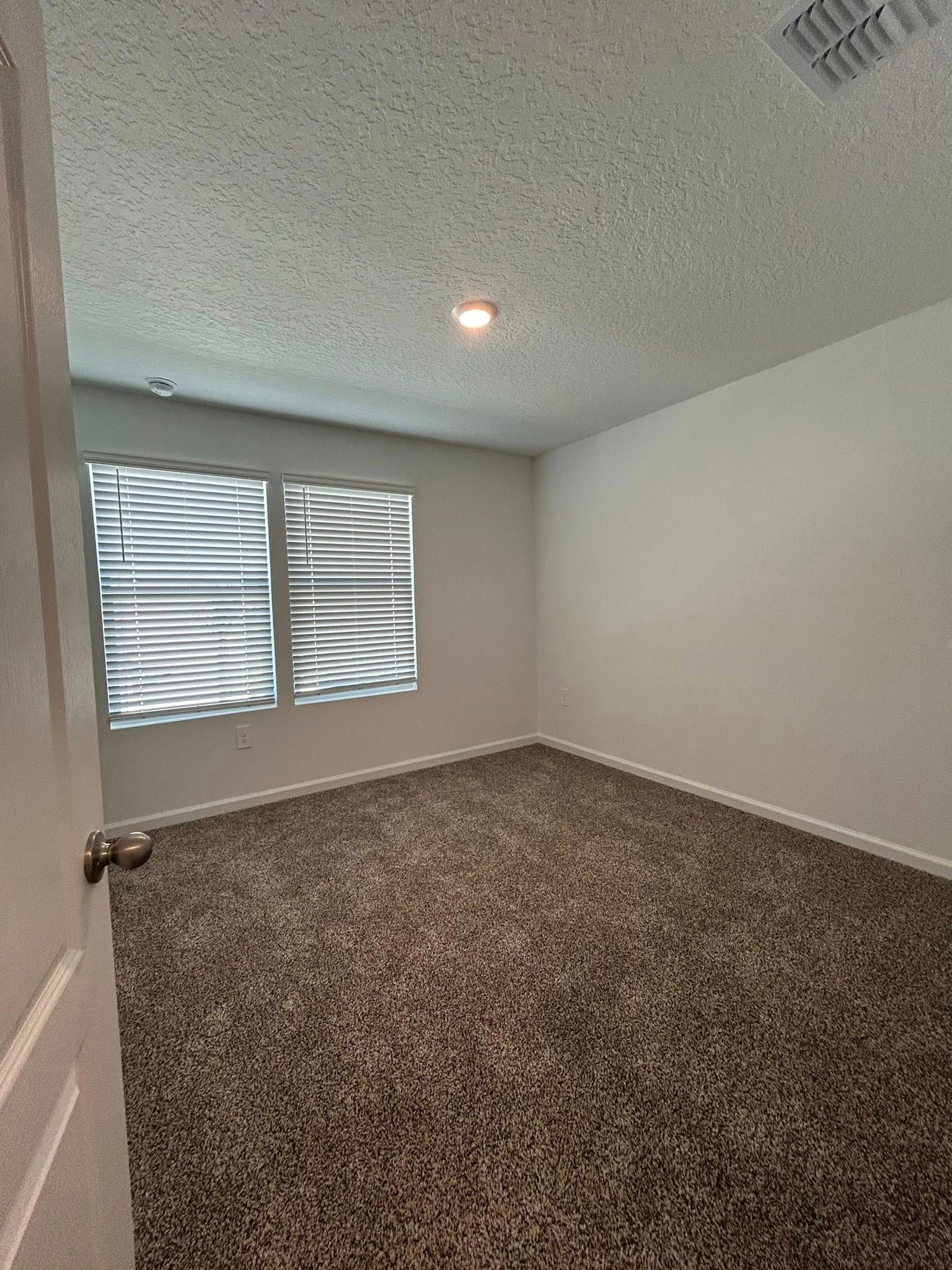 Empty room with brown carpet, white walls, two windows with blinds, and a door.