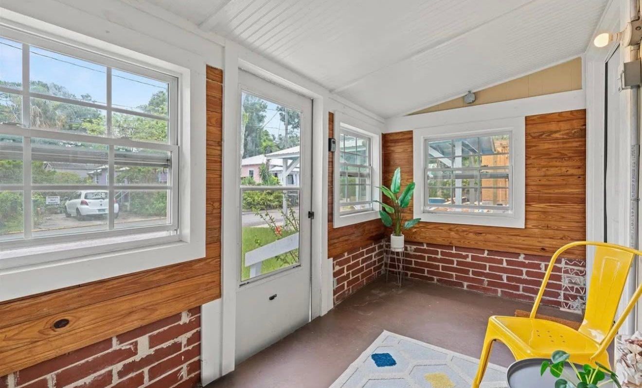 Sunroom with brick and wood walls, white windows and door, yellow chair, and a rug.