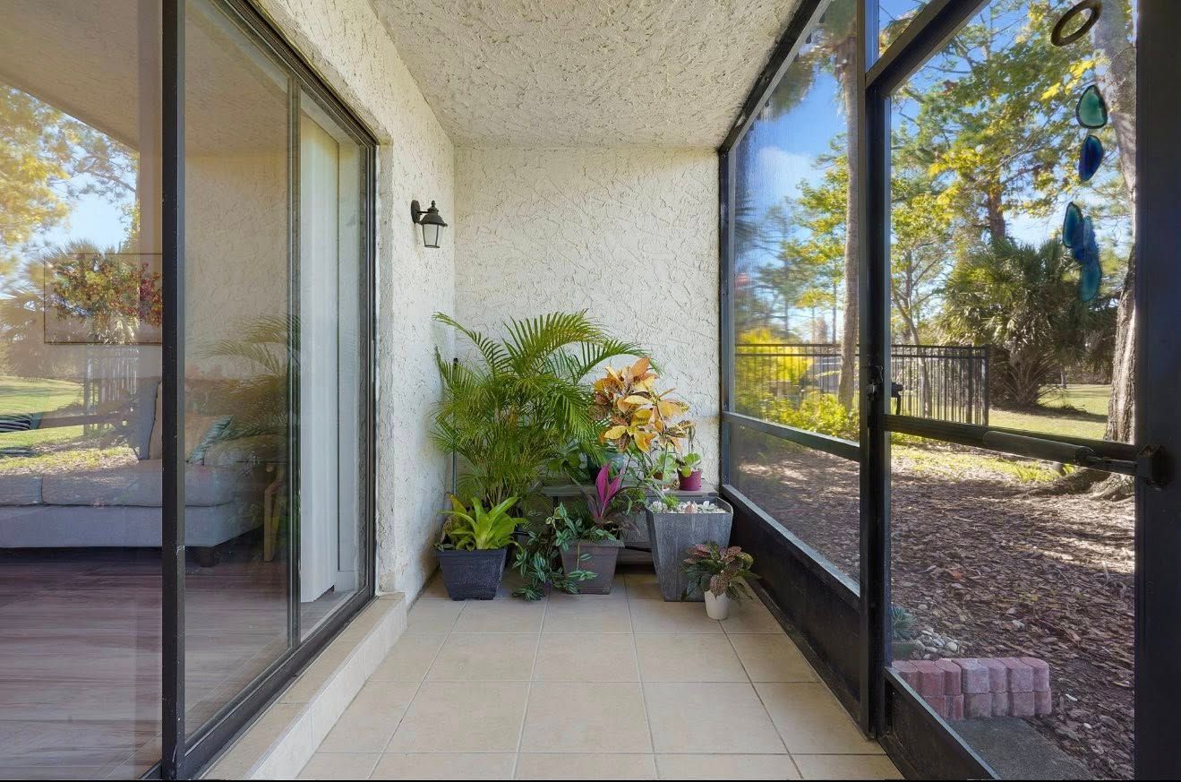Enclosed balcony with plants, beige walls, and large windows overlooking a yard with trees.
