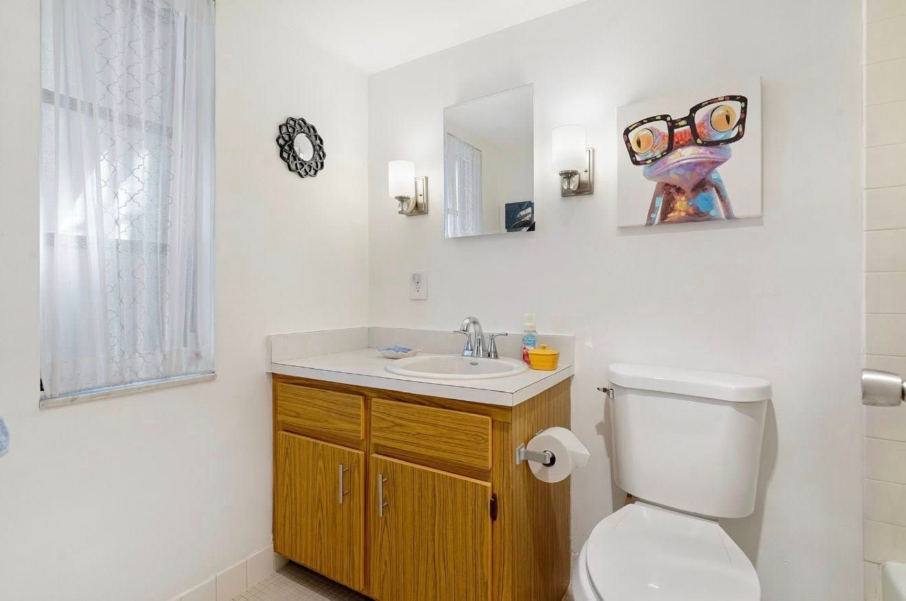 Bathroom with a vanity, toilet, and artwork; white walls, light wood cabinets, window with sheer curtain.