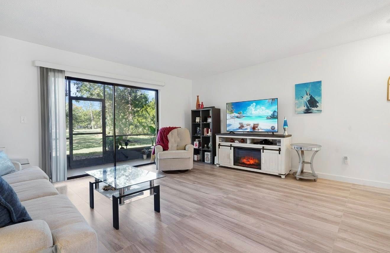 Living room with tan wood flooring, white walls, large window, and light-colored furniture.