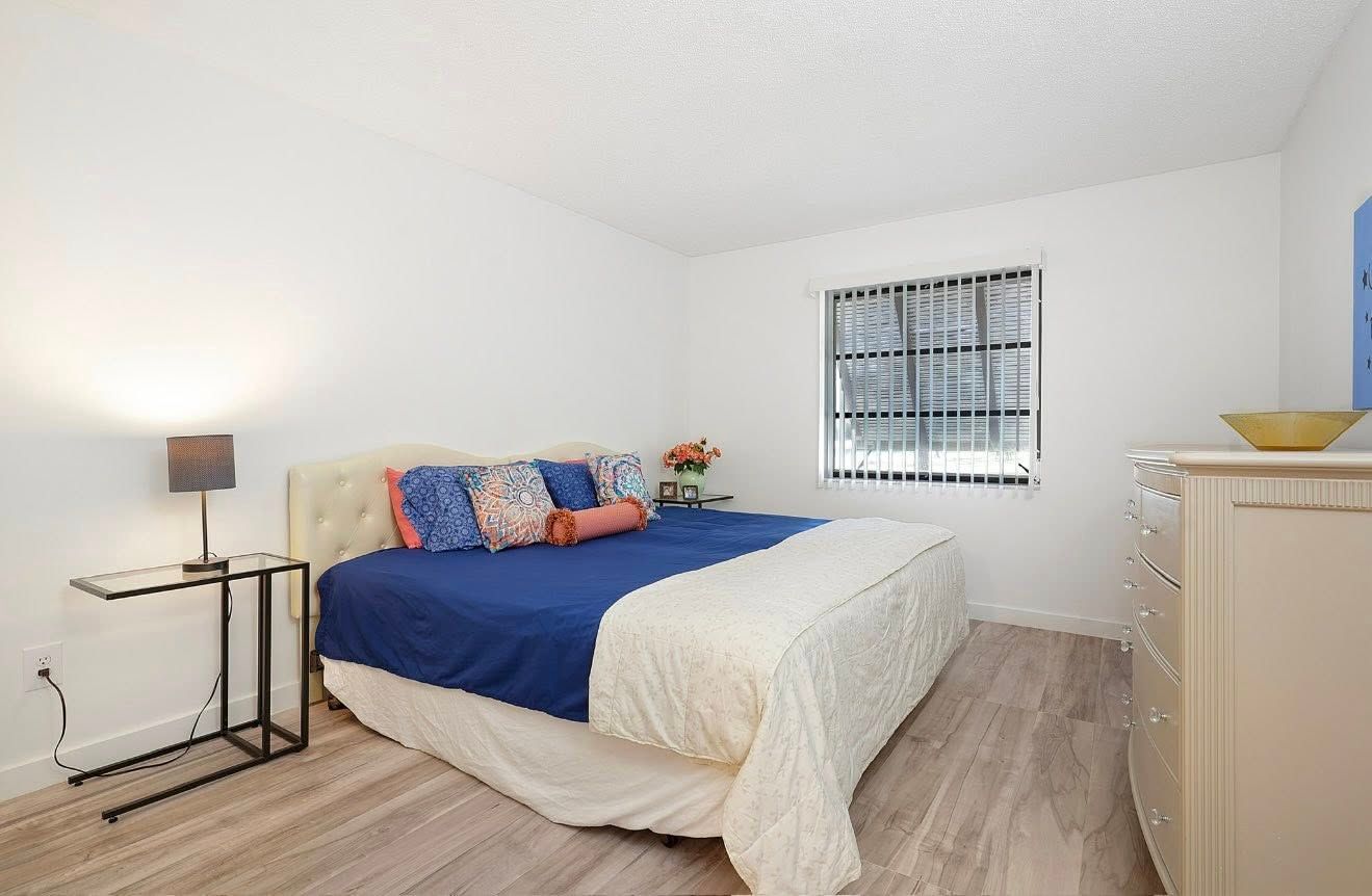 Bedroom with a bed, nightstand, dresser, and a window. White walls, wooden floor, blue and beige bedding.