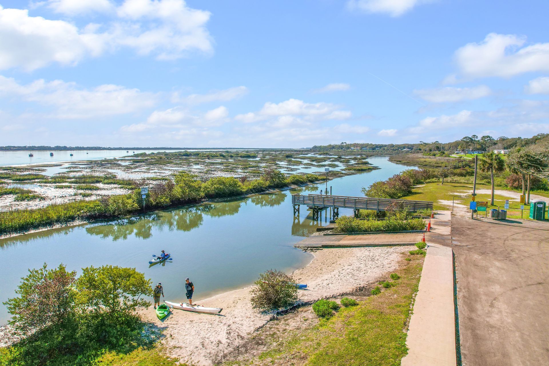 People prepare kayaks on a sandy beach beside a narrow waterway, with a wooden pier and wetlands under a blue, cloudy sky.
