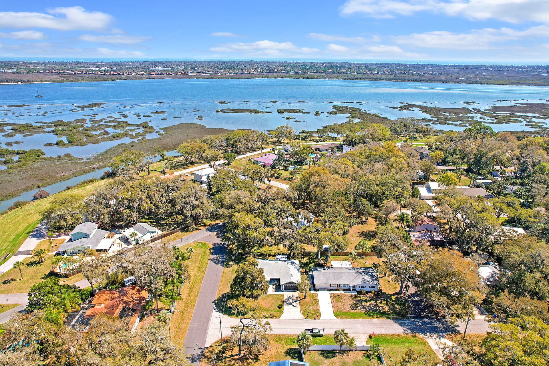 Aerial view of a coastal suburban neighborhood with houses nestled among trees, overlooking a blue bay and wetlands.