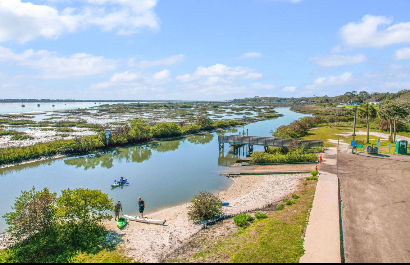 An aerial view of a calm waterway, featuring a small sandy beach, a wooden pier, and individuals launching kayaks.