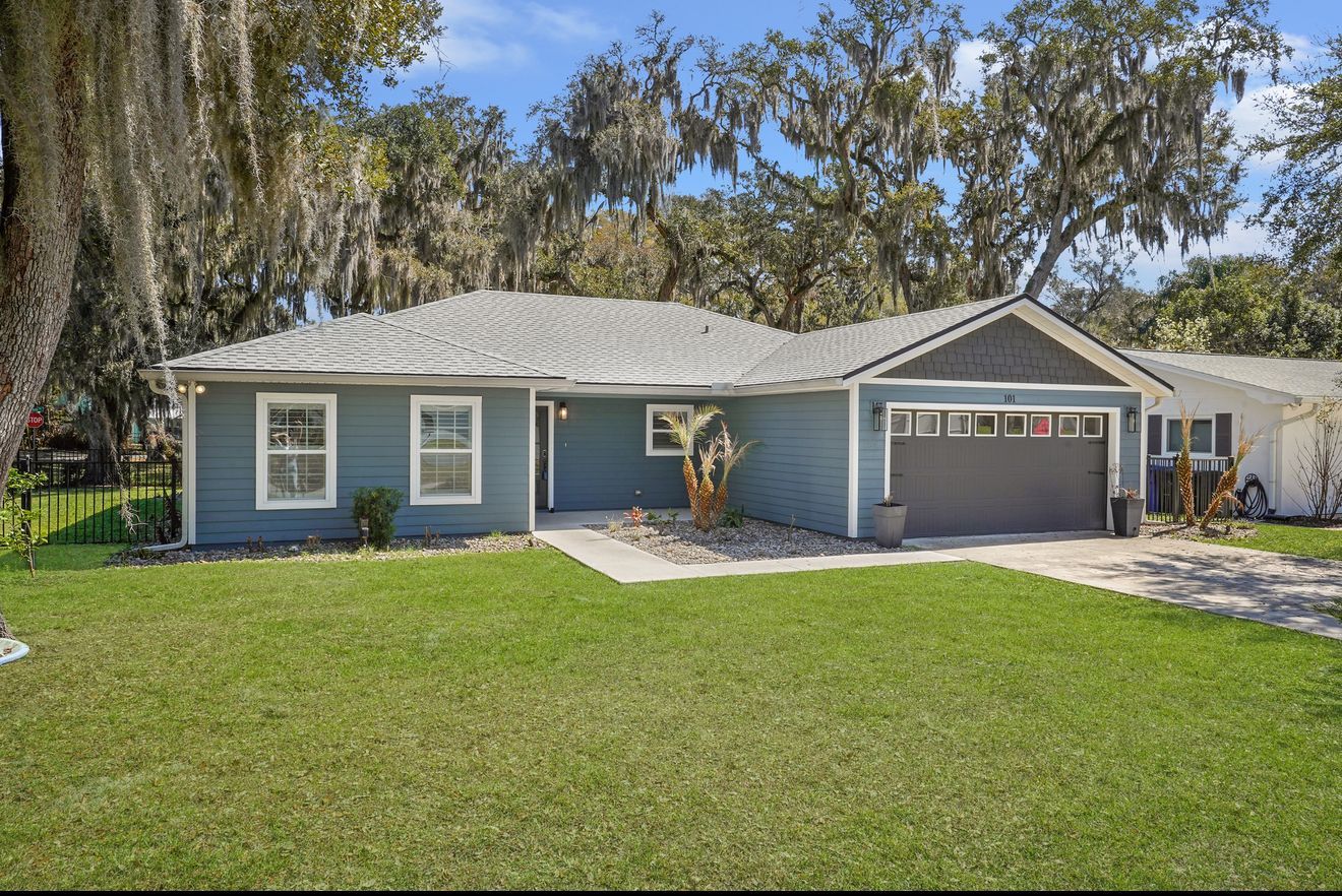 A single-story blue house with a grey roof, garage, and front lawn under trees with hanging moss on a sunny day.