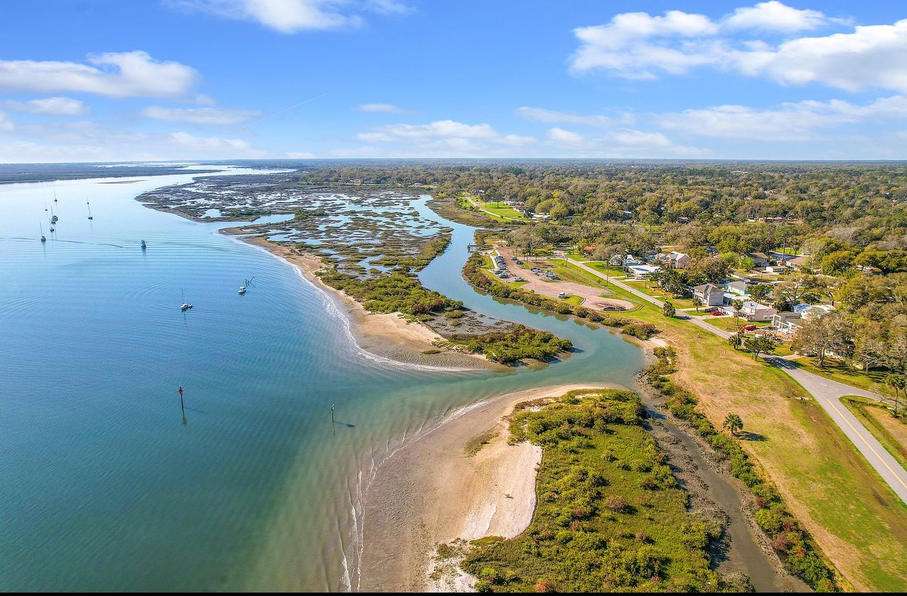 Aerial view of a coastal inlet where a narrow waterway meets a large, calm body of water near a tree-lined residential area.