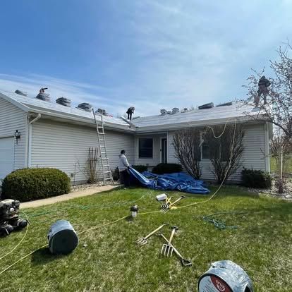 Roofers on a house under a blue sky. People are on the roof and around the home, with tools and a tarp in the yard.
