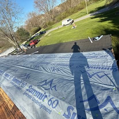 Person's shadow on a roof with underlayment, cars on a grassy hill.