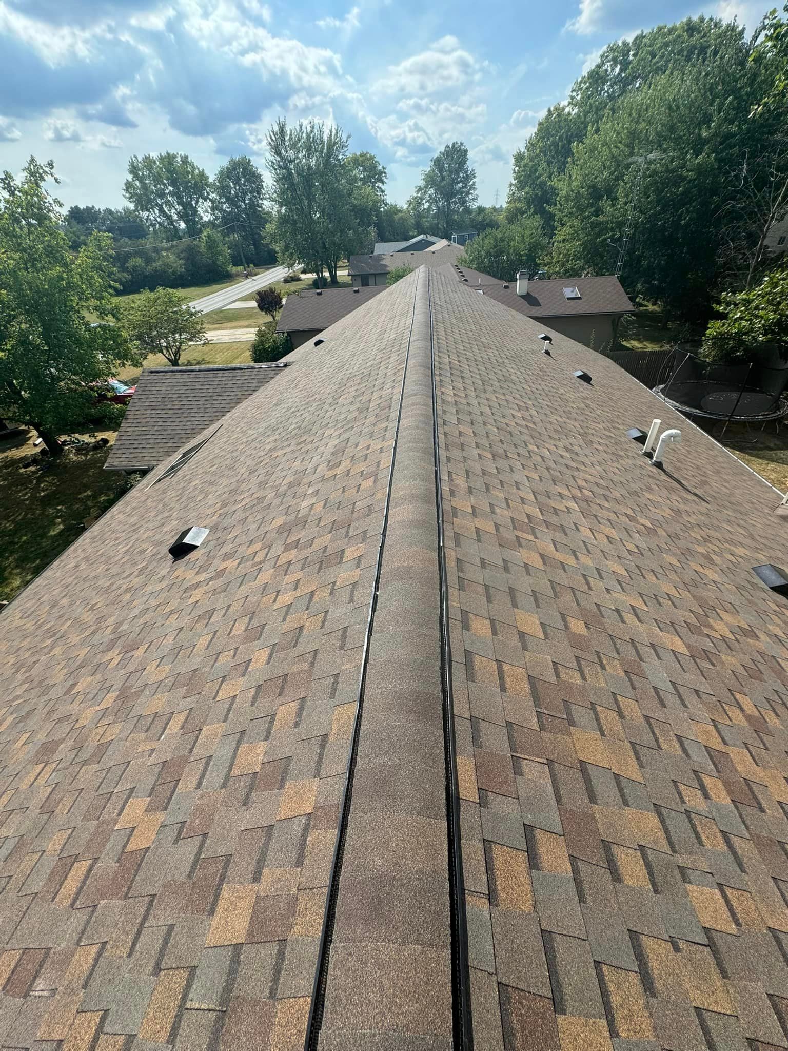 View of a rooftop with weathered shingles, trees and a street in the background under a blue sky.