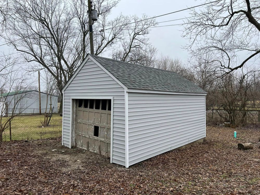 Garage with gray siding and a brown garage door in a yard with bare trees.