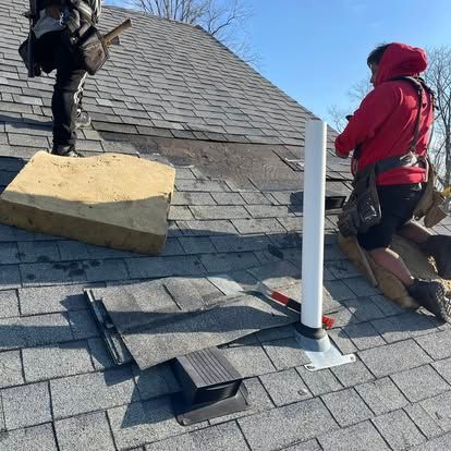 Two roofers repair damaged roof shingles on a sunny day. One in red, one in black, on a shingled roof.