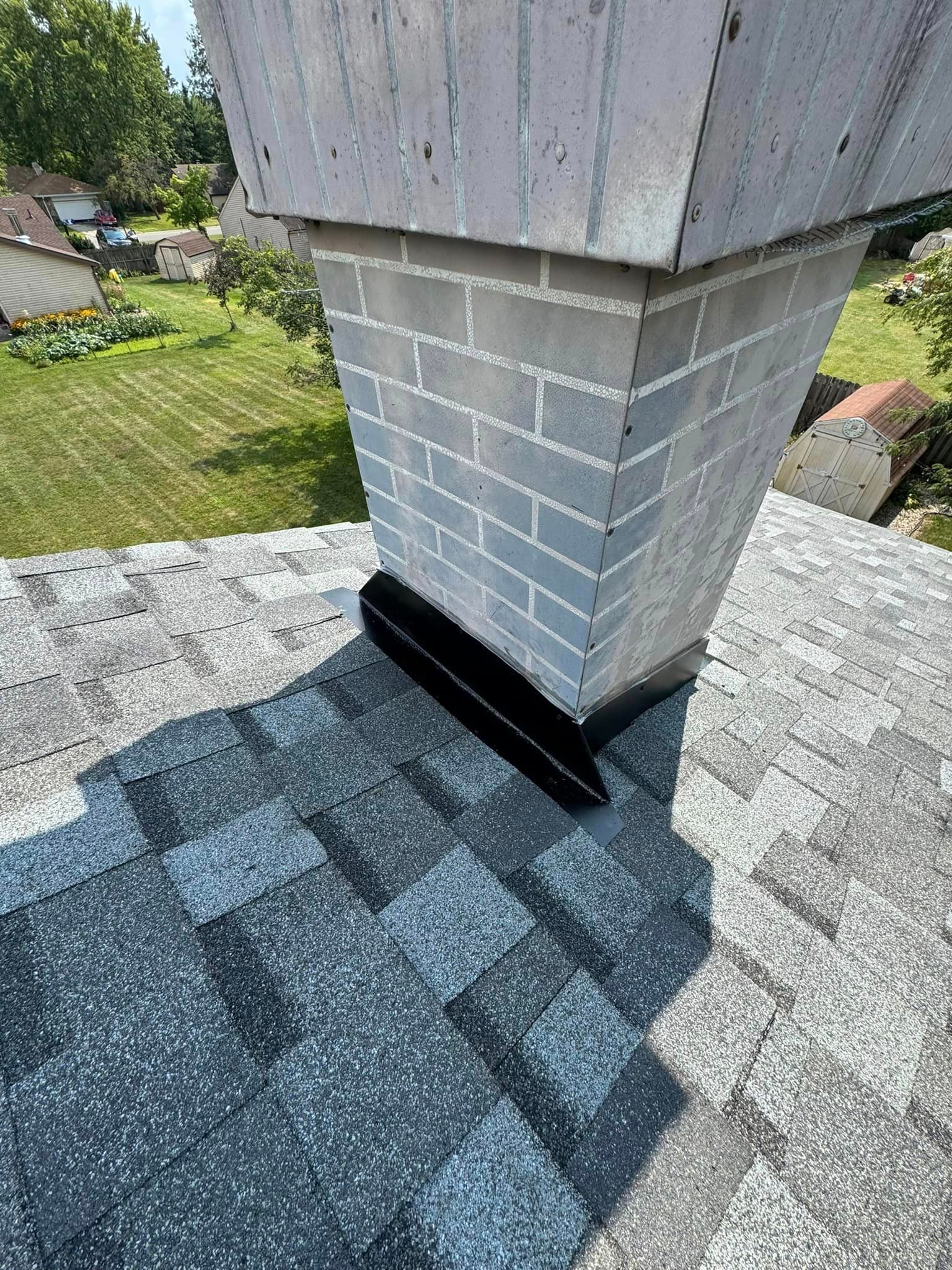 Chimney on a gray asphalt shingle roof with black flashing; sunny outdoor setting.