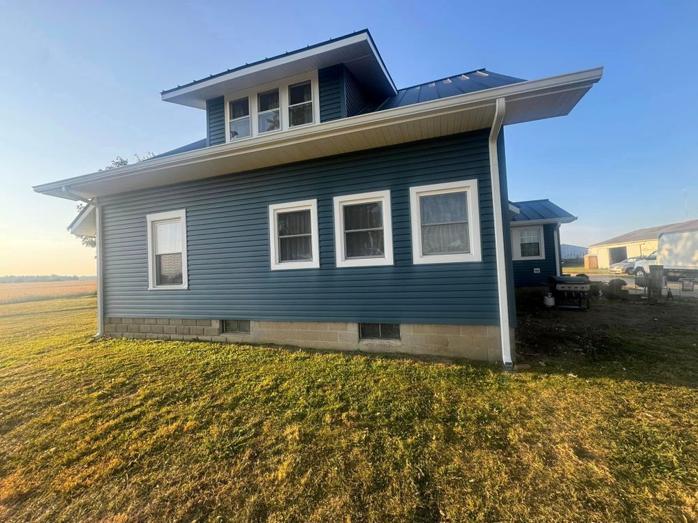 Blue-sided house with white trim, windows, and a dark roof, set in a grassy field under a blue sky.