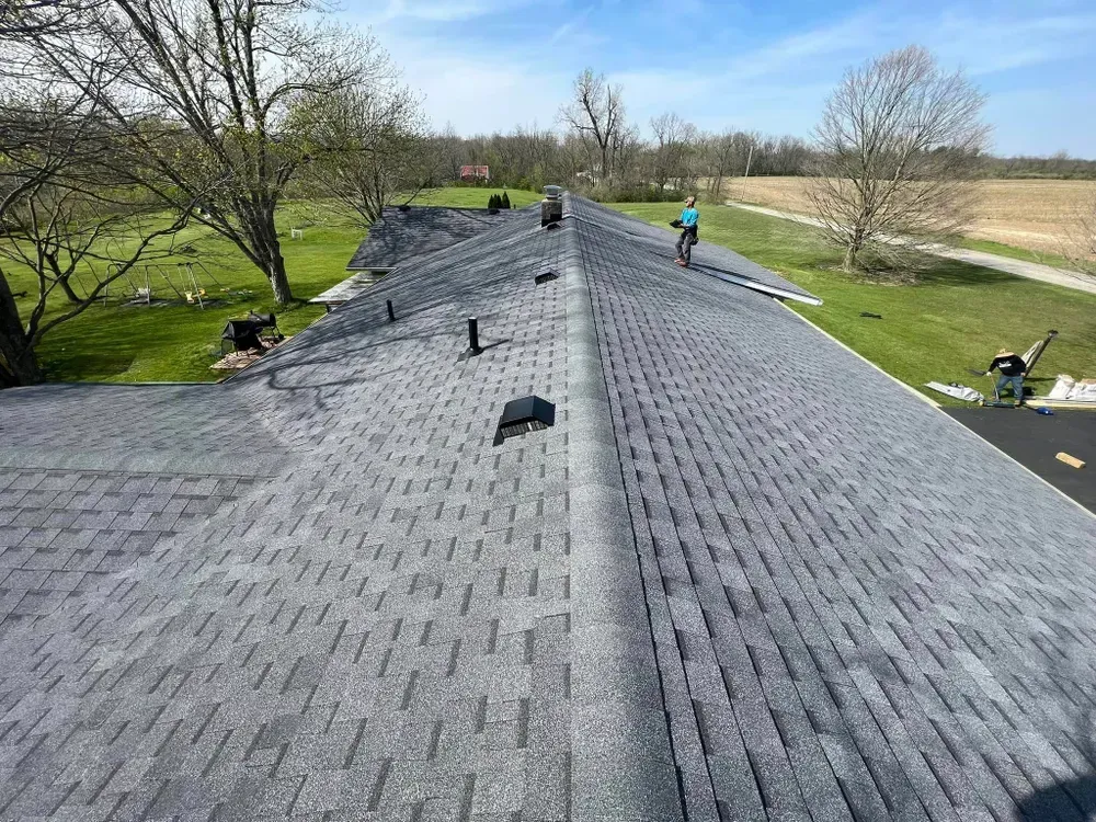 Roofers installing new asphalt shingles on a house roof, set in a rural landscape under a blue sky.