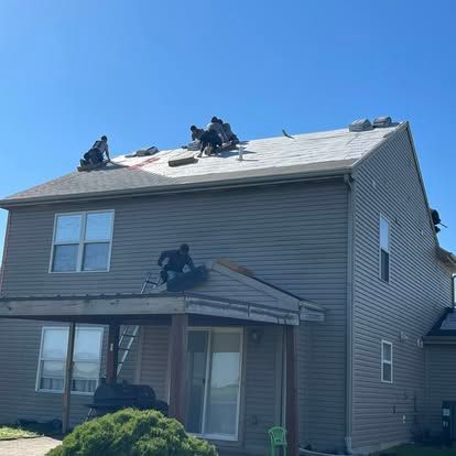 Roofers working on a two-story house with gray siding under a clear, blue sky.