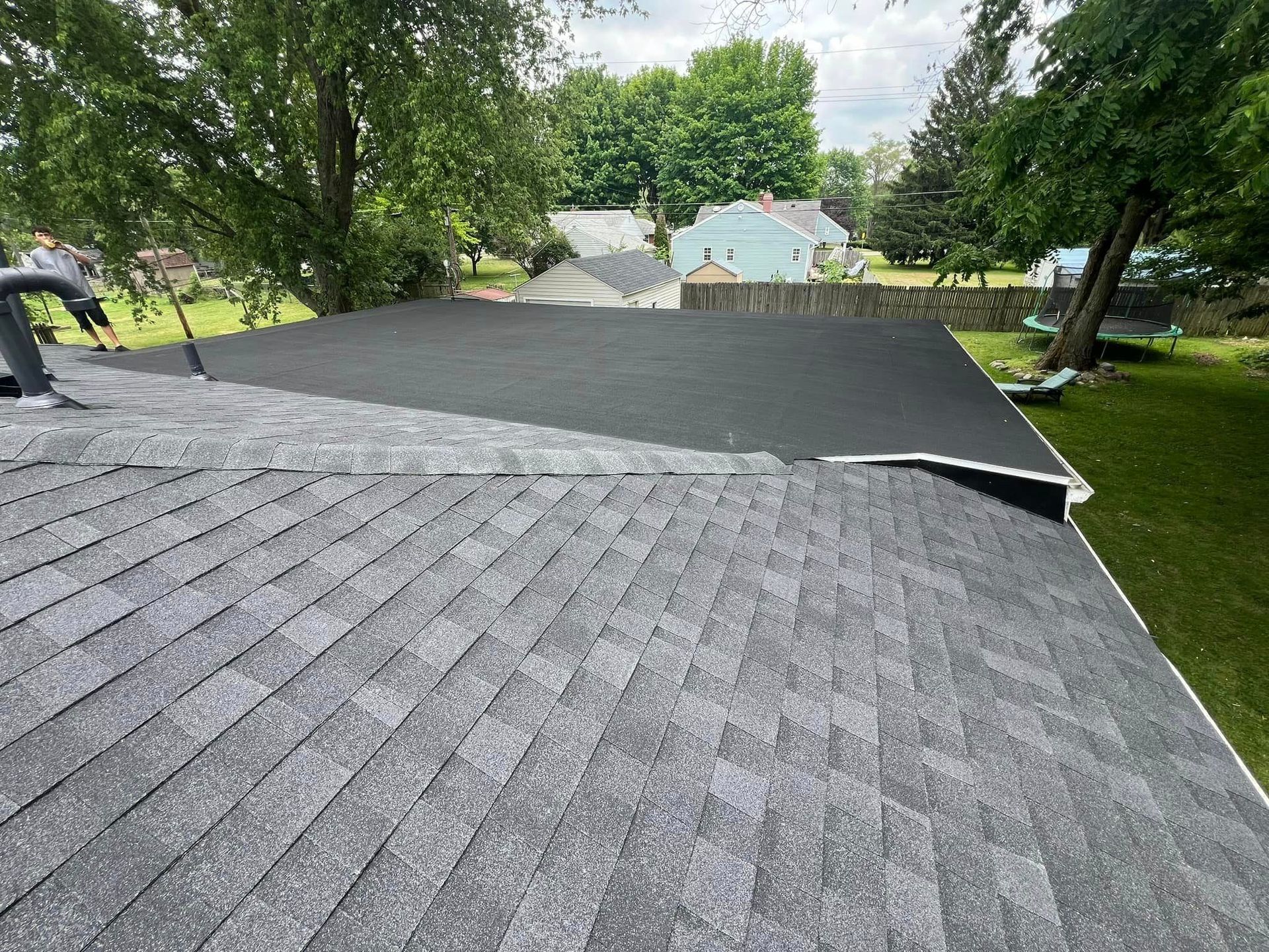 Roof partially covered with gray shingles, black material on top, surrounded by trees and houses.