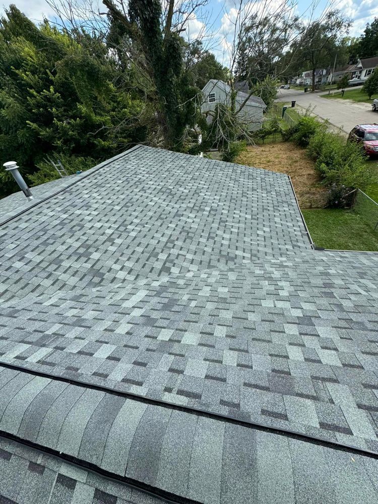 Gray asphalt shingle roof on a residential house with trees and houses in the background under a sunny sky.