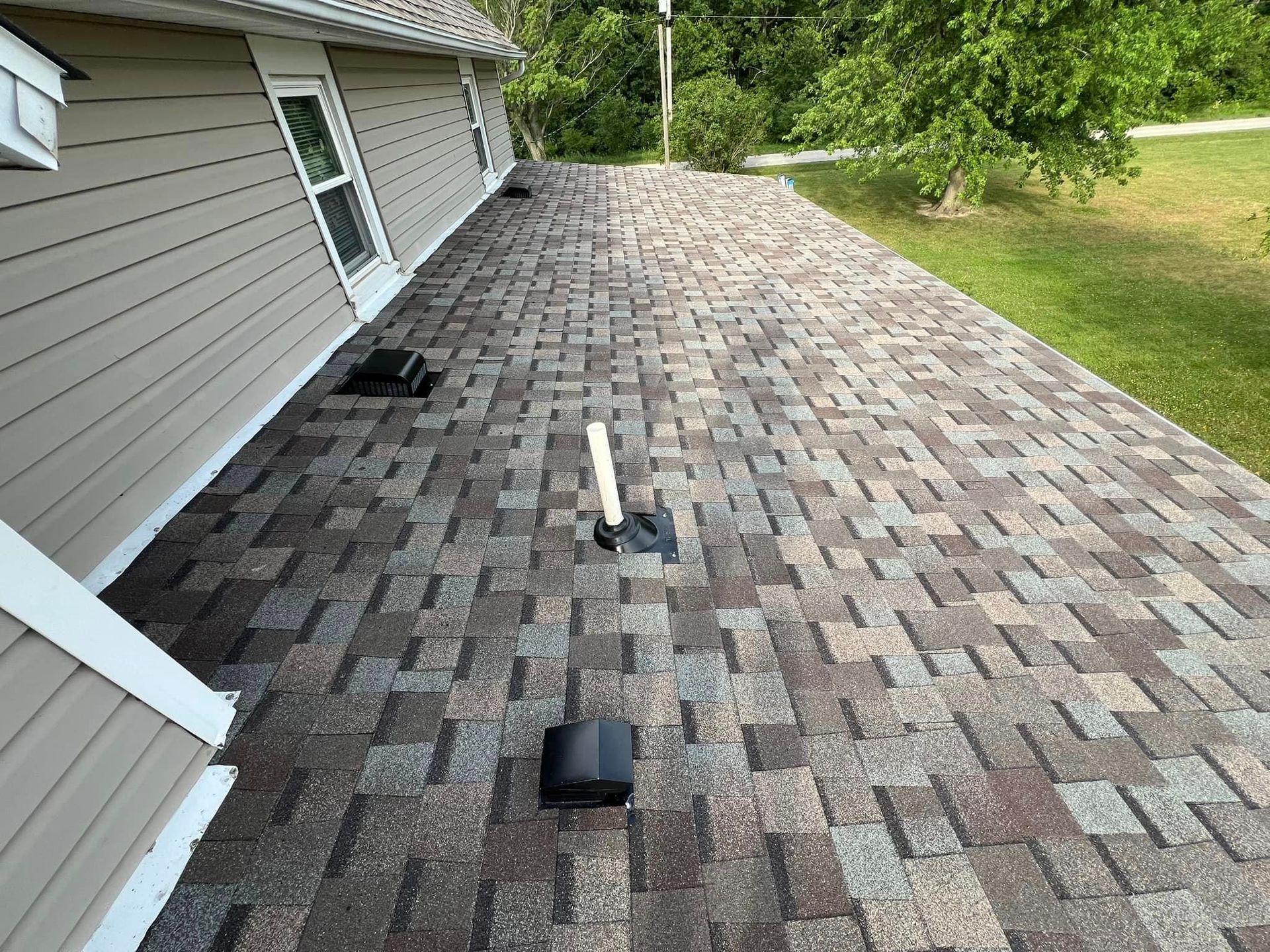 A low-angle view of a brown shingled roof with vents, next to a light-colored house, green grass, and trees.