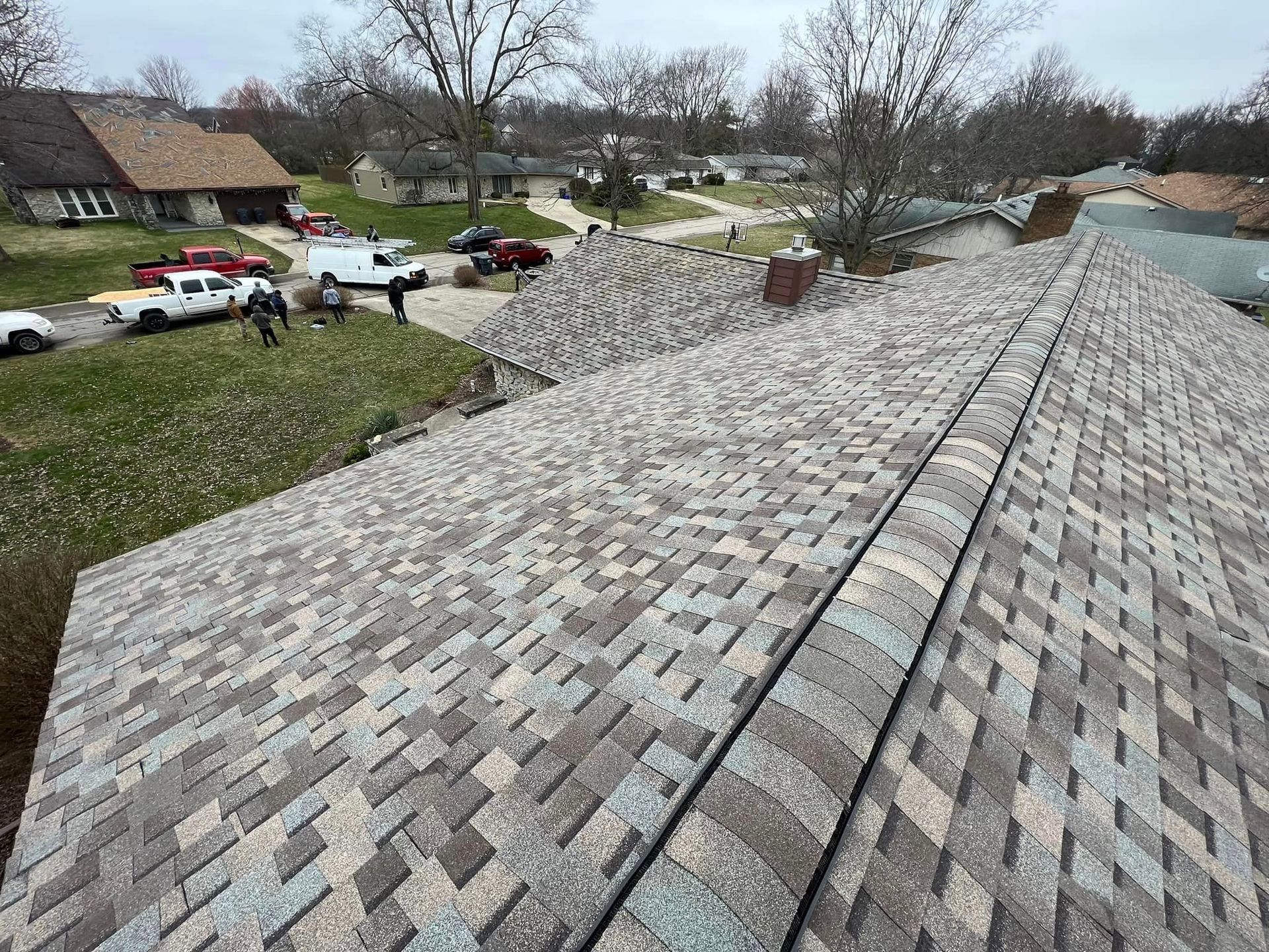 Newly shingled roof with varied brown, gray, and blue tones; workers and vehicles on adjacent lawn.