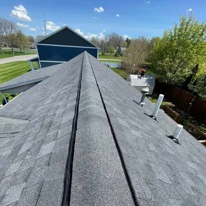 Gray shingled rooftop with a central ridge, chimneys, and vents, on a sunny day.