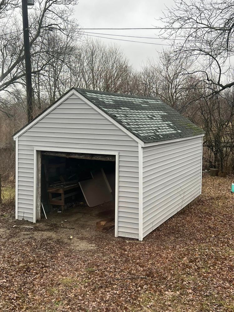 Gray, single-car garage with open door, siding, and weathered roof. Surrounded by trees and brown leaves.