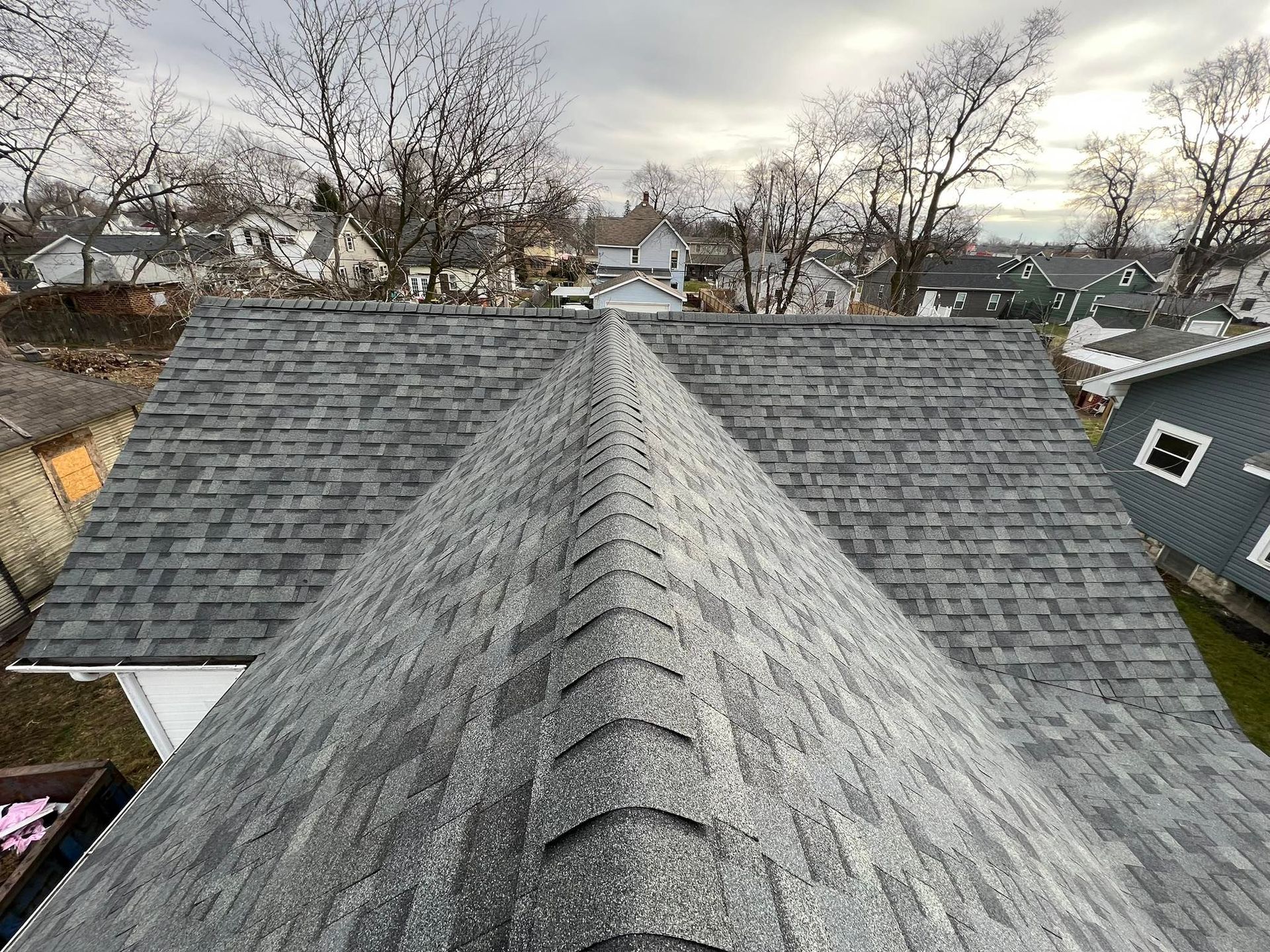 Gray shingled roof from a high angle view, with neighborhood houses in the background on a cloudy day.