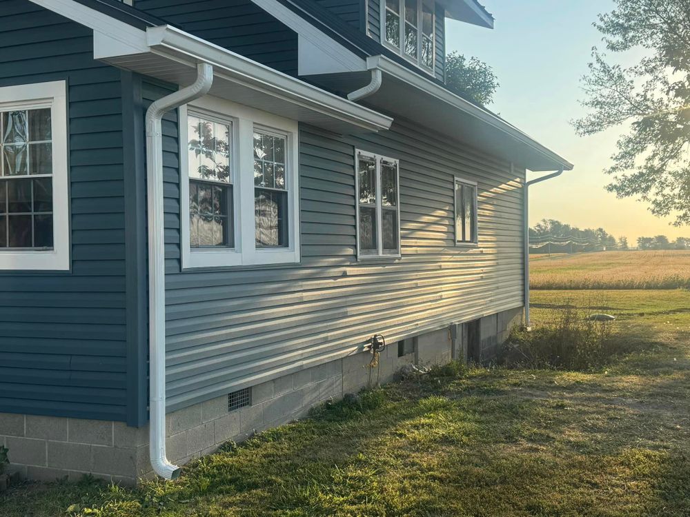 Blue house with white trim, windows, and gutters; side view with a field in the background.