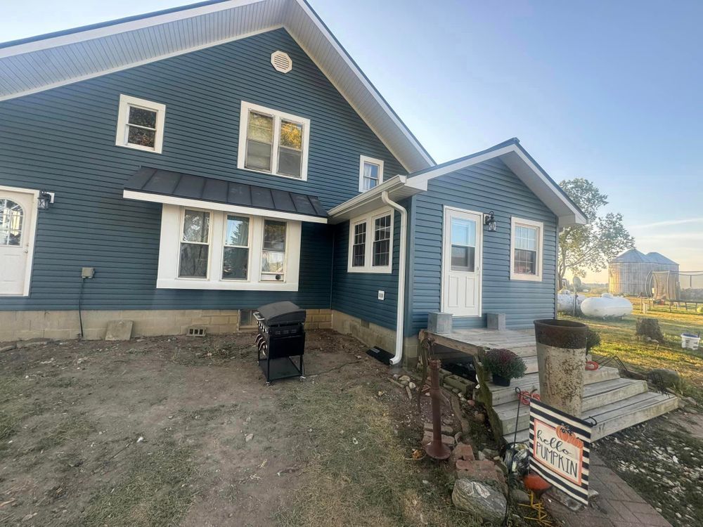 Blue house with white trim, black awning, front door, and steps; setting is rural, exterior.