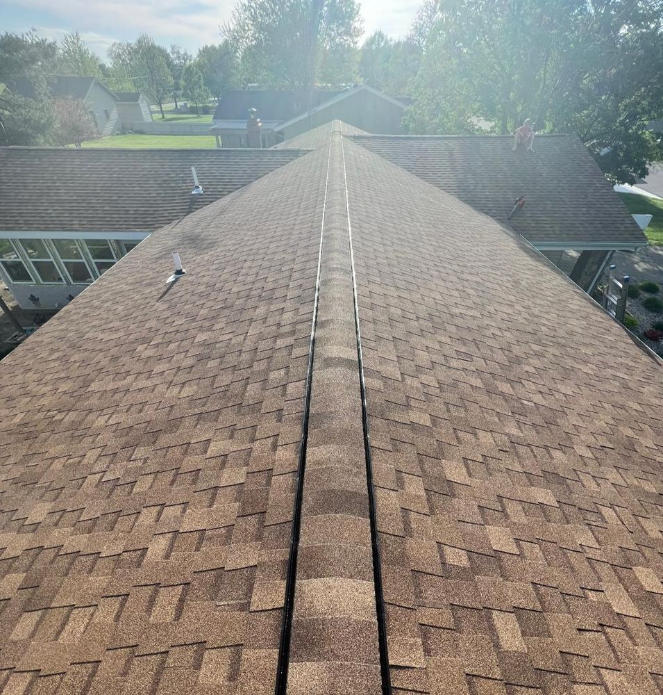 A high-angle view of a brown shingled roof, with a vent, trees, and other houses visible.