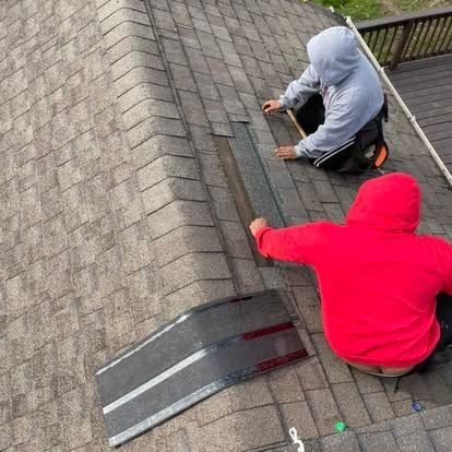 Two roofers in hoodies repair a shingle seam on a roof, one with a tool.