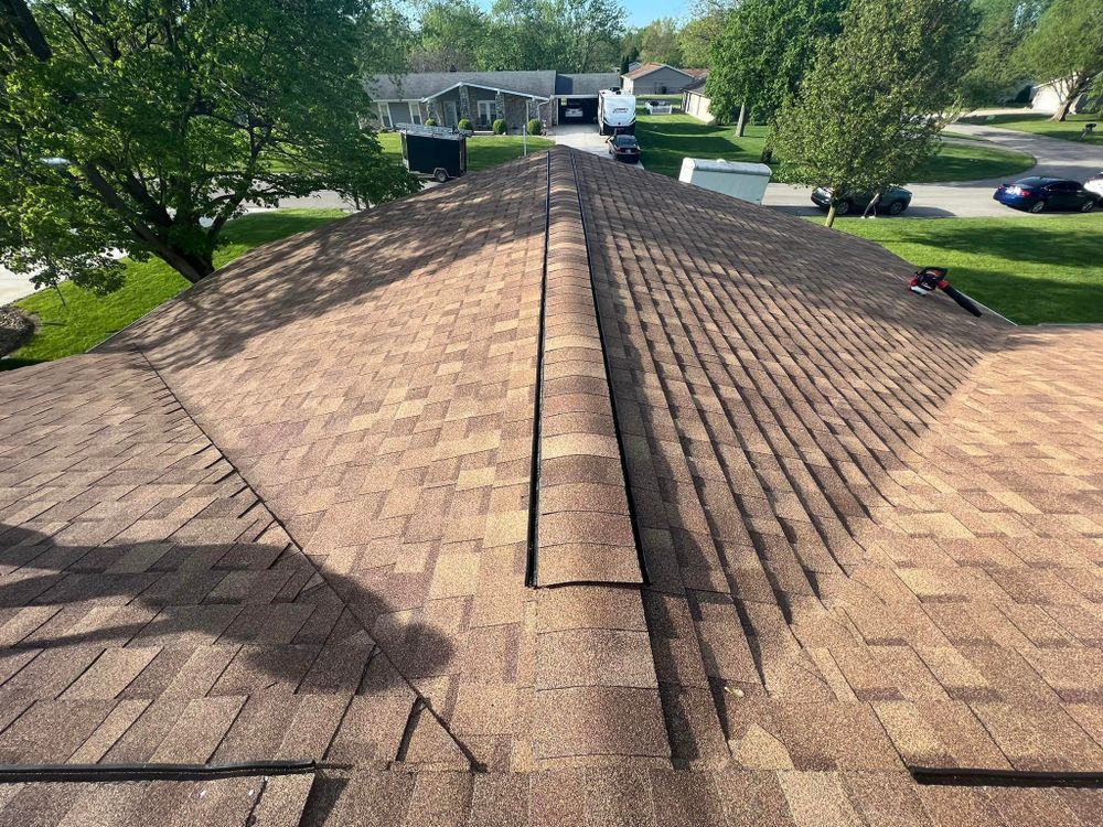 Brown shingle roof with a ridge vent, viewed from above; trees and houses in the background.