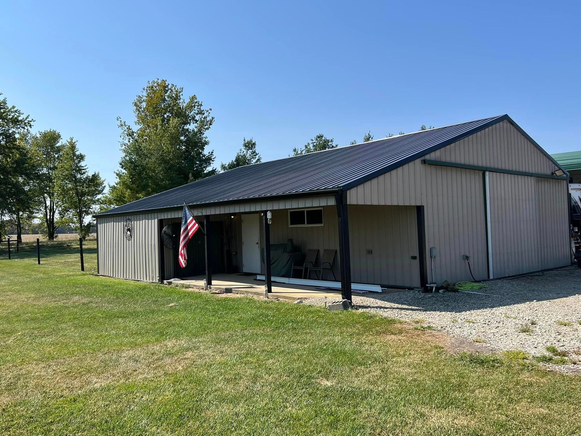 Tan barn with black roof and porch, American flag, green grass.