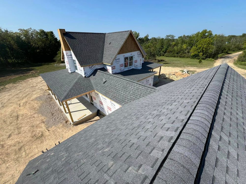House under construction, gray shingle roof, paper-wrapped siding, open land, blue sky.