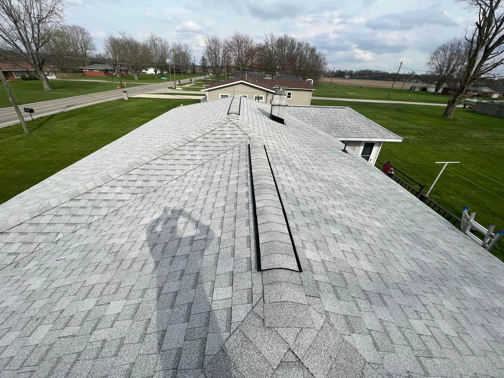 Overhead view of a light gray shingled roof with a damaged section and the surrounding green yard and houses.