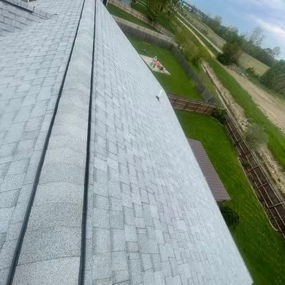 Gray shingled roof of a house, angled view. Green lawn, fence, and dirt path in the background.