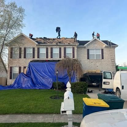 Roofers working on a two-story house with a tarp covering the front; recycling bins in the foreground.