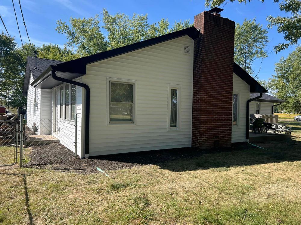 White house with black trim, brick chimney, and black gutters. Green lawn and trees in background.
