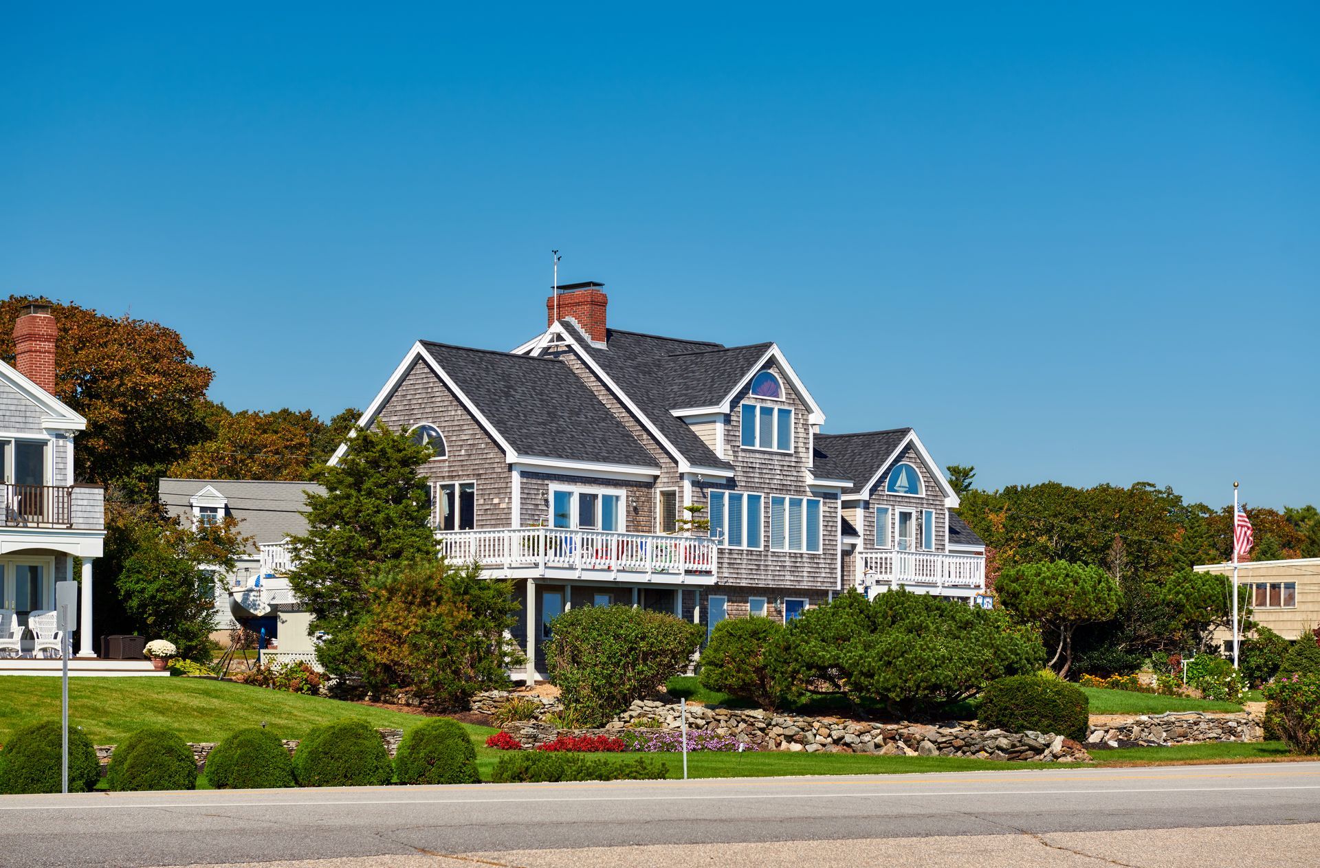 Two-story gray-shingled house with blue shutters, balconies, and a dark roof, under a blue sky.