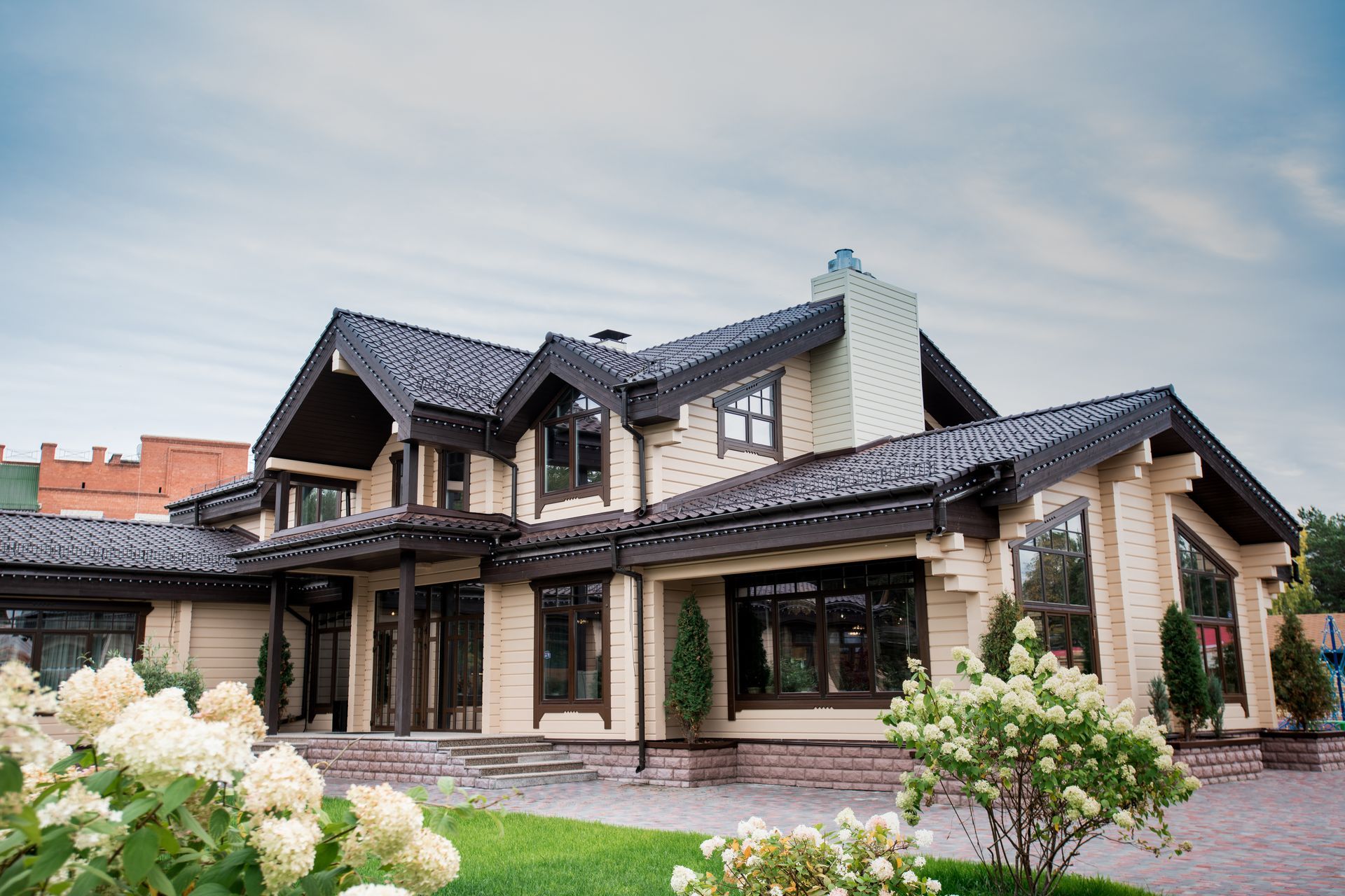 Wooden house with a dark roof and beige siding, surrounded by landscaping and hydrangeas, set under a cloudy sky.
